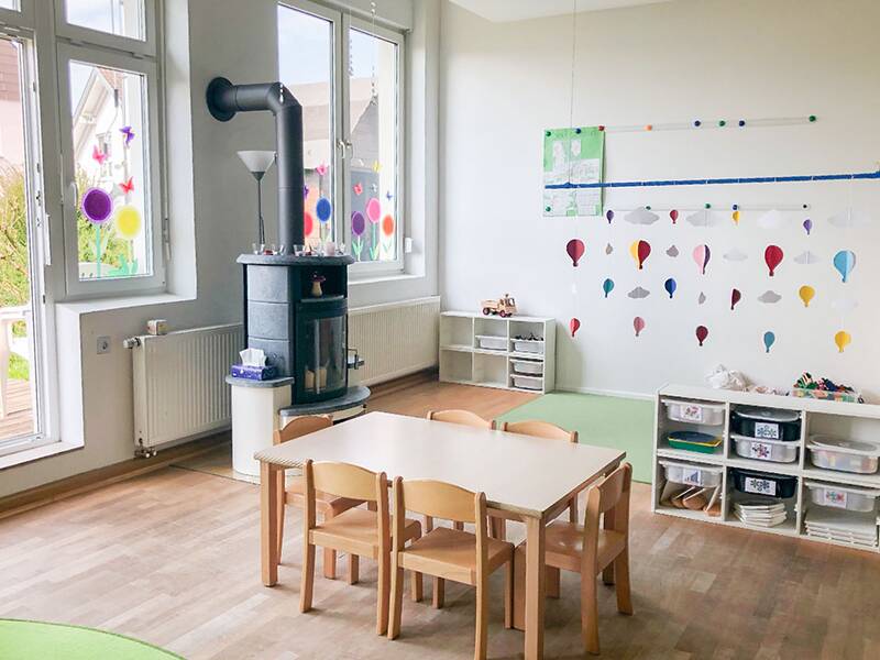 Ein heller, gemütlicher Raum mit einem Holztisch und Stühlen, einem Kaminofen, bunten Fenstermalereien und Wanddekorationen in Form von Heißluftballons.