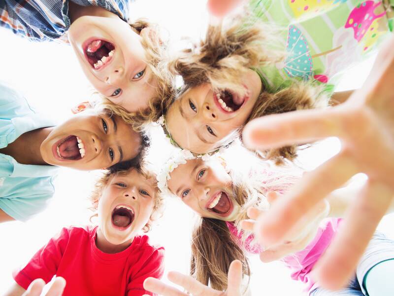 Fünf lachende Kinder blicken fröhlich von oben in die Kamera und strecken die Hände aus.