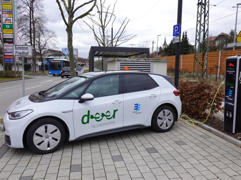 Ein weißes Elektroauto mit der Aufschrift "deer" steht an einer Ladestation auf einem Parkplatz in einer städtischen Umgebung.