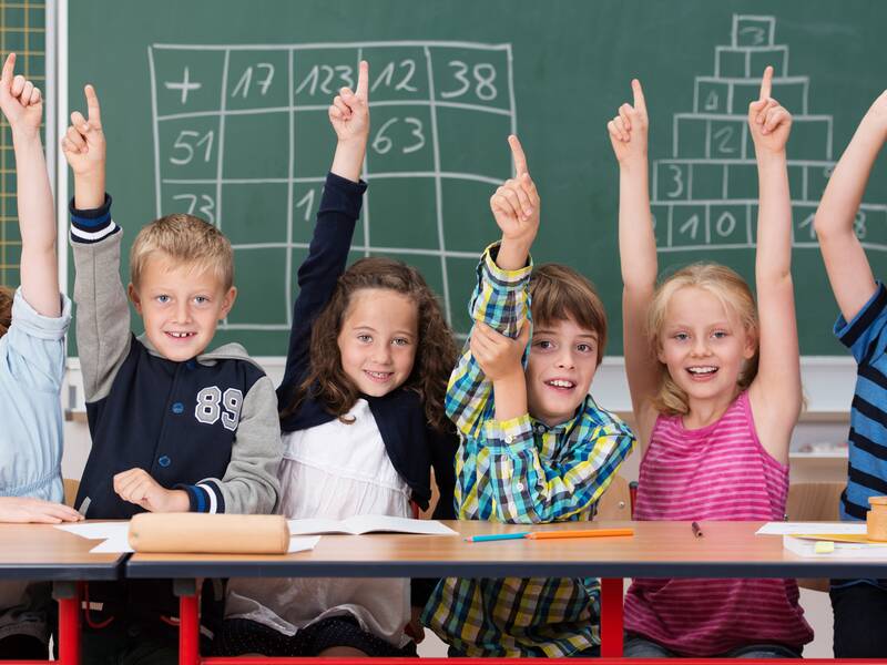 Sechs Kinder sitzen in einer Schulklasse vor einer Tafel und melden sich. Auf der Tafel stehen Mathematikaufgaben für Grundschulkinder