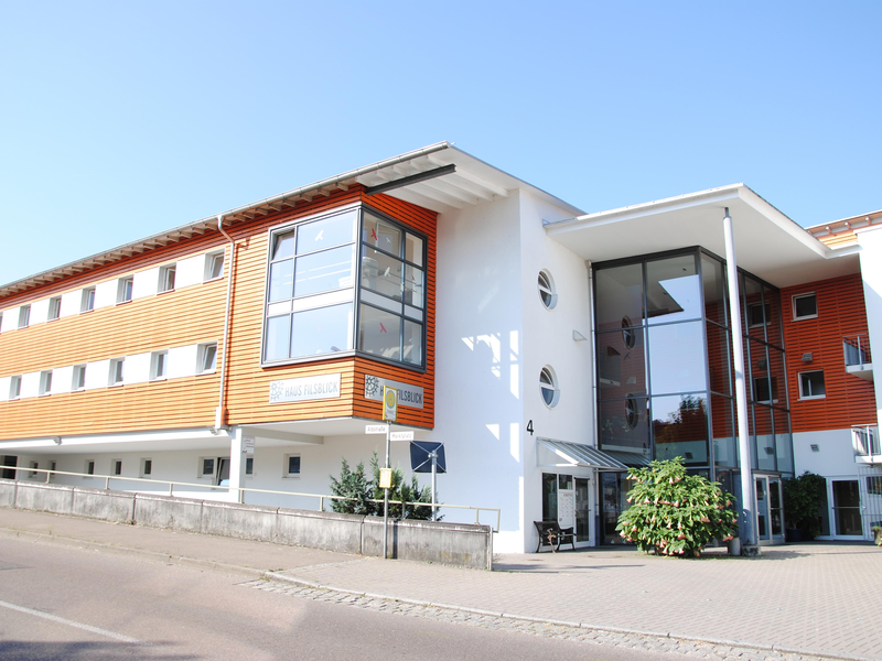 Das Haus Filsblick in Ebersbach von außen. Ein modernes Gebäude mit einer Holzfassade, großen Glasfenstern und einem flachen Dach, umgeben von einem gepflasterten Bereich und einer Straße.